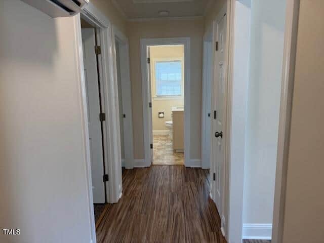 A hallway with wood flooring leads to a bathroom at the end. The walls are painted light beige. Doors on the left and right give access to other rooms. The bathroom features a window with blinds and visible plumbing fixtures.