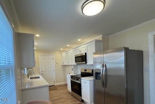Modern kitchen with white cabinets, stainless steel appliances including a refrigerator and microwave, and a gas stove. A bright overhead light and recessed lighting illuminate the space. There is a window with blinds to the left.