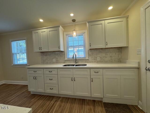 A kitchen with white cabinetry and a light-colored tile backsplash. There is a window above a stainless steel sink and faucet. Recessed lighting and a pendant light illuminate the space. The floor is wood, and the walls are beige.
