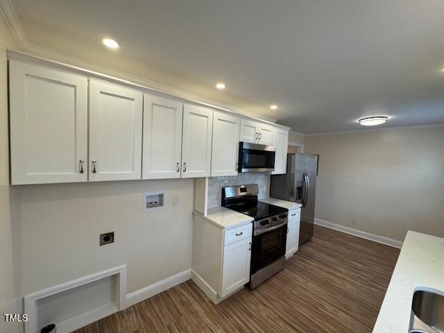 A modern kitchen with white cabinets, a stainless steel microwave, oven, and refrigerator. The backsplash is light-colored tile, and the floor is wood. There is a blank space for a washer and dryer on the left wall.