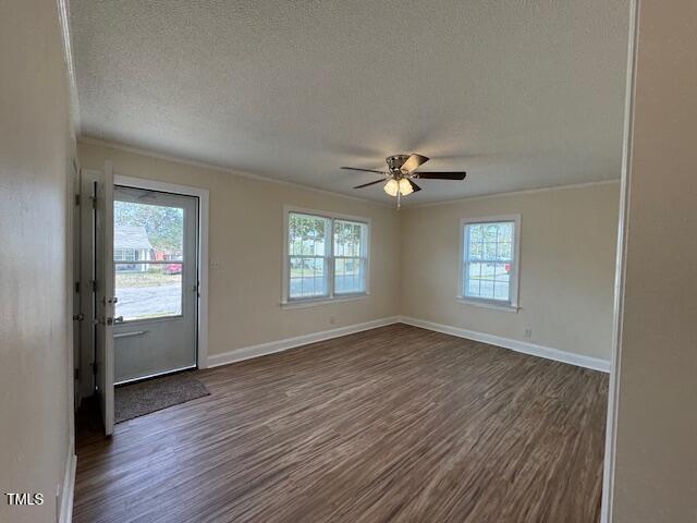 A vacant room with beige walls and wood flooring. It has a ceiling fan with lights, three windows providing natural light, and a glass door leading outside. The space is empty, with no furniture or decorations.