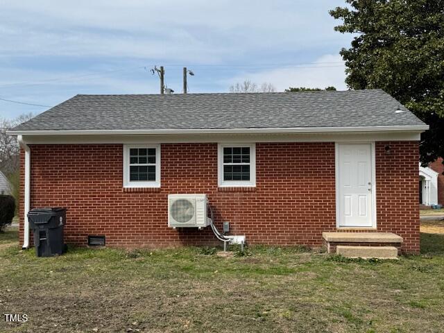 A small red brick house with a gray shingle roof, featuring three windows and a white door. An air conditioning unit is mounted on the exterior wall. Two trash bins are placed on the left side. The lawn in front is sparse and patchy.