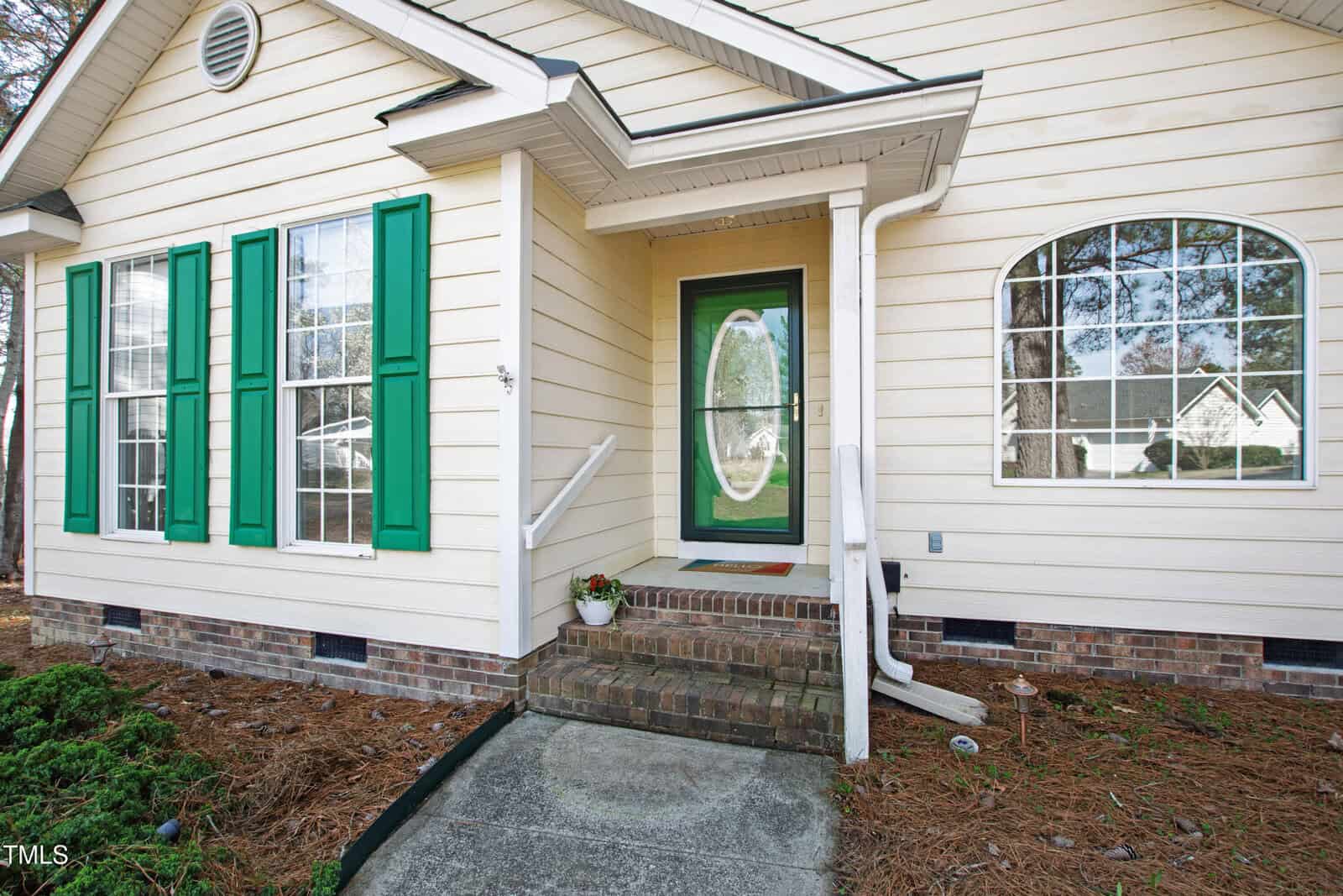 The image shows the front of a beige house with green shutters and a green front door with an oval window. Two steps lead up to the door, and there are arched windows beside it. A downspout is visible, and the ground is covered with mulch.