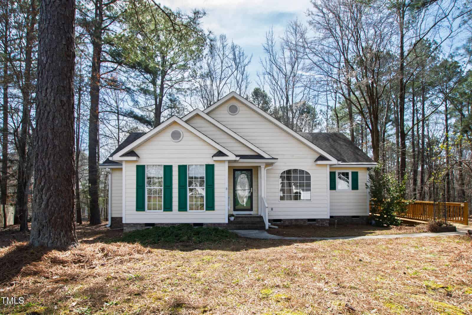 A small, single-story house with white siding and green shutters is nestled among the tall trees of Creedmoor. Located at 2776 Jordan Court, its front features a gabled roof and a cozy set of stairs leading to the entrance, while a wooden deck extends invitingly from one side.