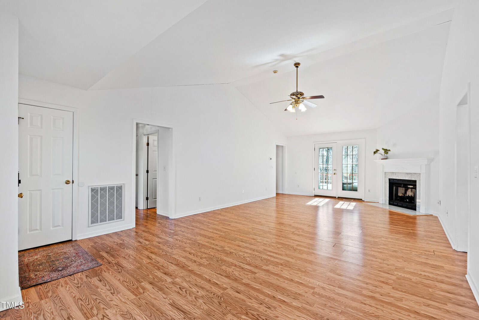 Spacious empty living room with light wood flooring and white walls. A ceiling fan with light is centered above. There is a fireplace with a mantel on the right and French doors leading outside in the background. Doorways and vents are visible.