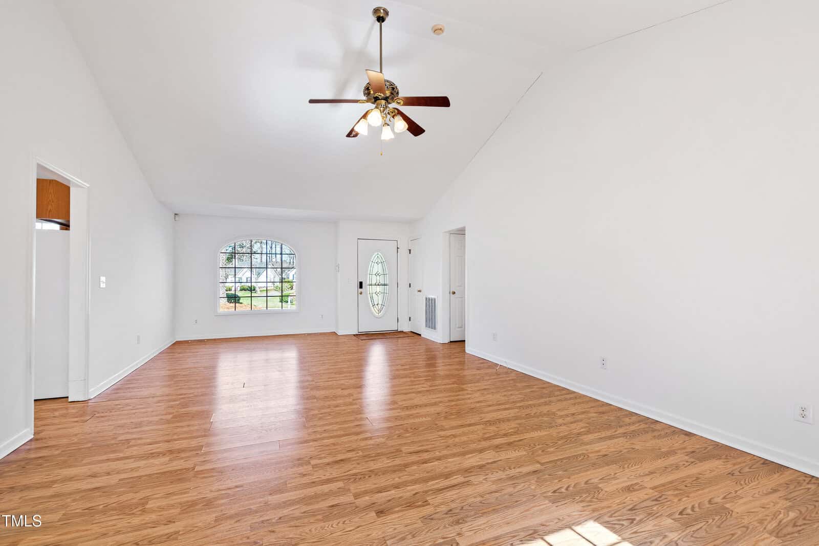 Spacious living room with light wood flooring, white walls, and a vaulted ceiling. A ceiling fan with lights hangs above. An arched window and a glass-paneled front door are on the far wall, allowing natural light to enter.
