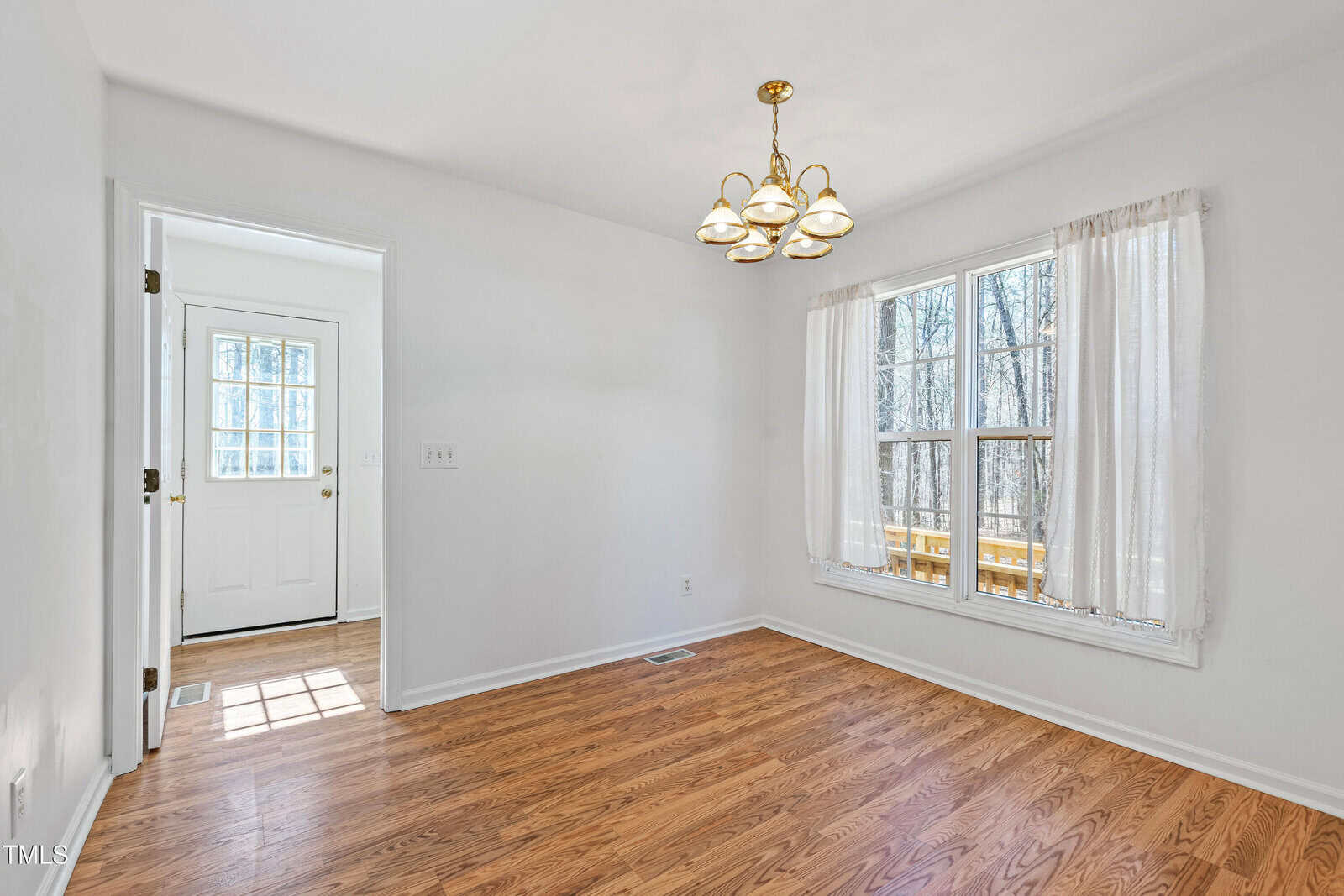 A bright dining room with hardwood floors, a five-light brass chandelier, and a window with sheer curtains overlooking a wooden deck. To the left is a white door with glass panels leading outside.