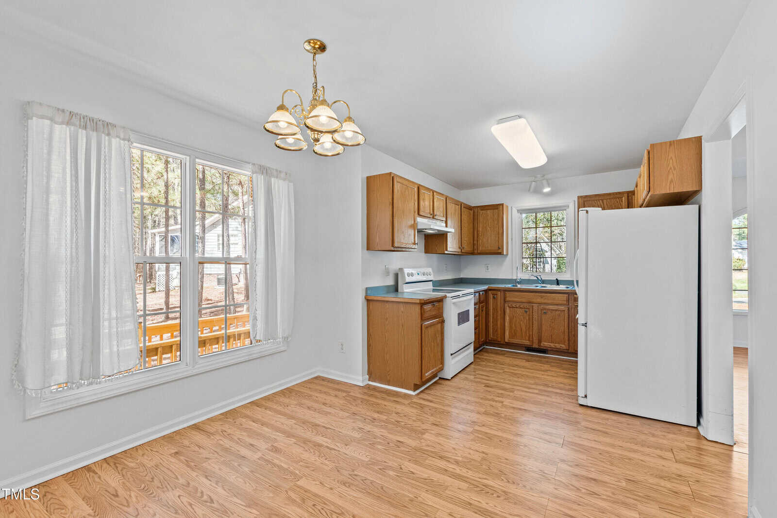 A kitchen with wooden cabinets, a white stove, and refrigerator. A dining area with hardwood floors is visible, featuring a window with sheer curtains and a chandelier above. The room is well-lit with natural light.