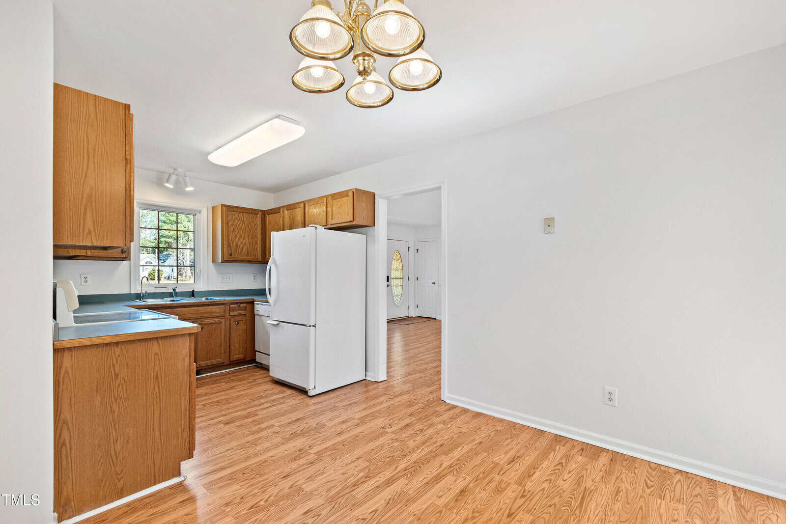 Kitchen and dining area with wooden cabinets, white appliances, and hardwood flooring. A chandelier hangs above the dining space. A window is above the kitchen sink, and a round front door is visible through an open doorway.
