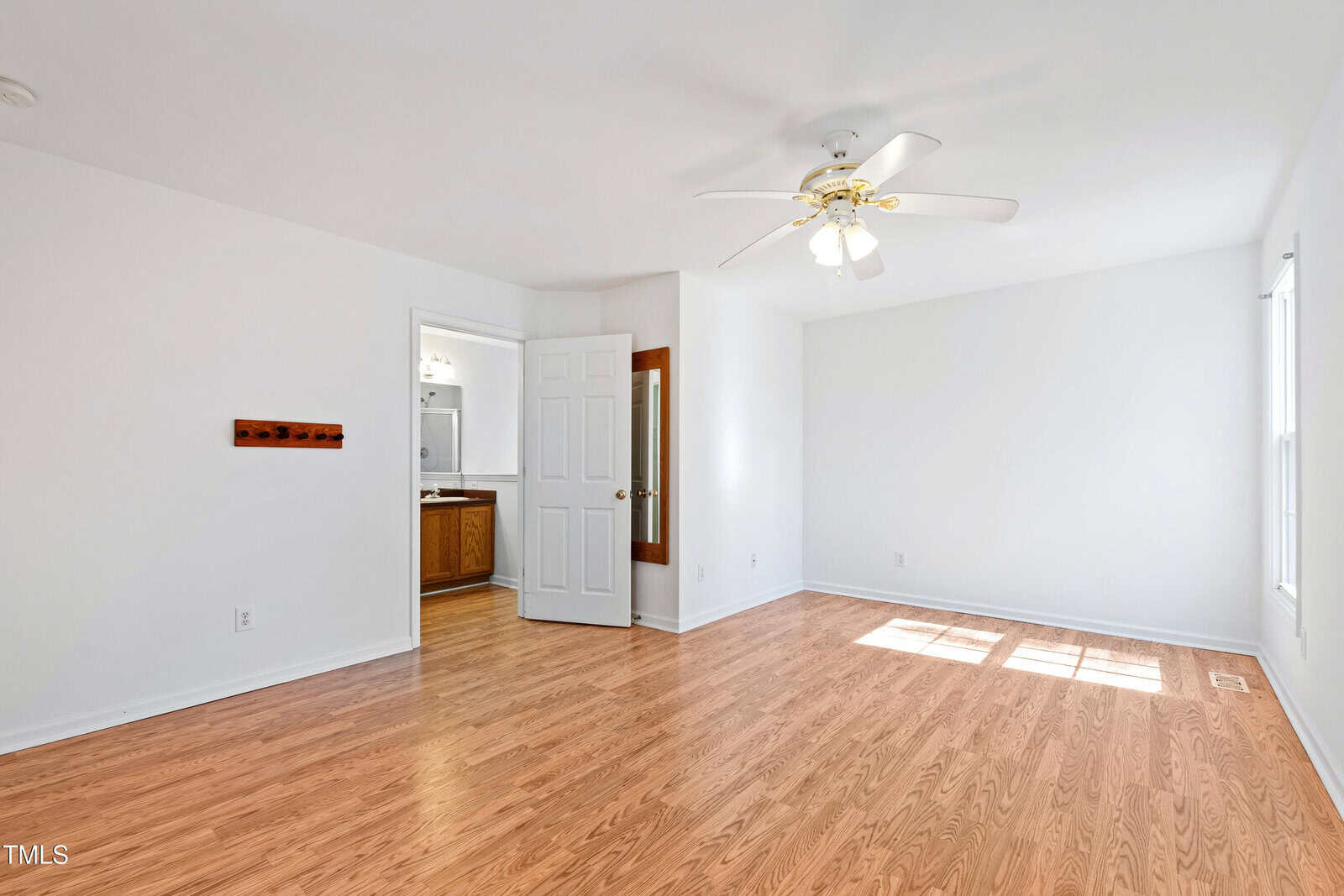 A bright, empty room with light wood flooring, white walls, and a ceiling fan with lights. A door is open, revealing a glimpse of a bathroom with a wooden cabinet and a sink. Sunlight streams in through a window on the right.
