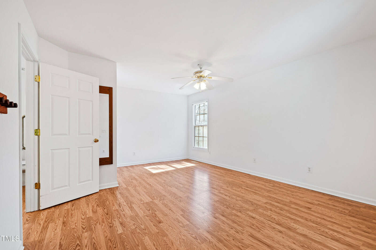 An empty room with light-colored hardwood flooring and white walls. A ceiling fan with lights is centered on the ceiling. A single window lets in natural light. An open door is visible on the left side.