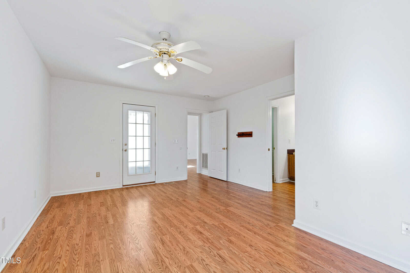 An empty room with white walls and wooden flooring. A ceiling fan with a light fixture is centered above. The room has a door with glass panels leading outside and two open doorways to other rooms.