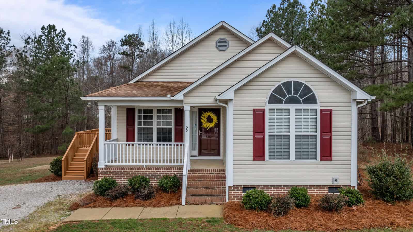 Single-story beige house with white trim, an arched front window, a covered porch with steps, a wooden side staircase, brown shutters, and landscaped shrubs set against a backdrop of trees and blue sky.