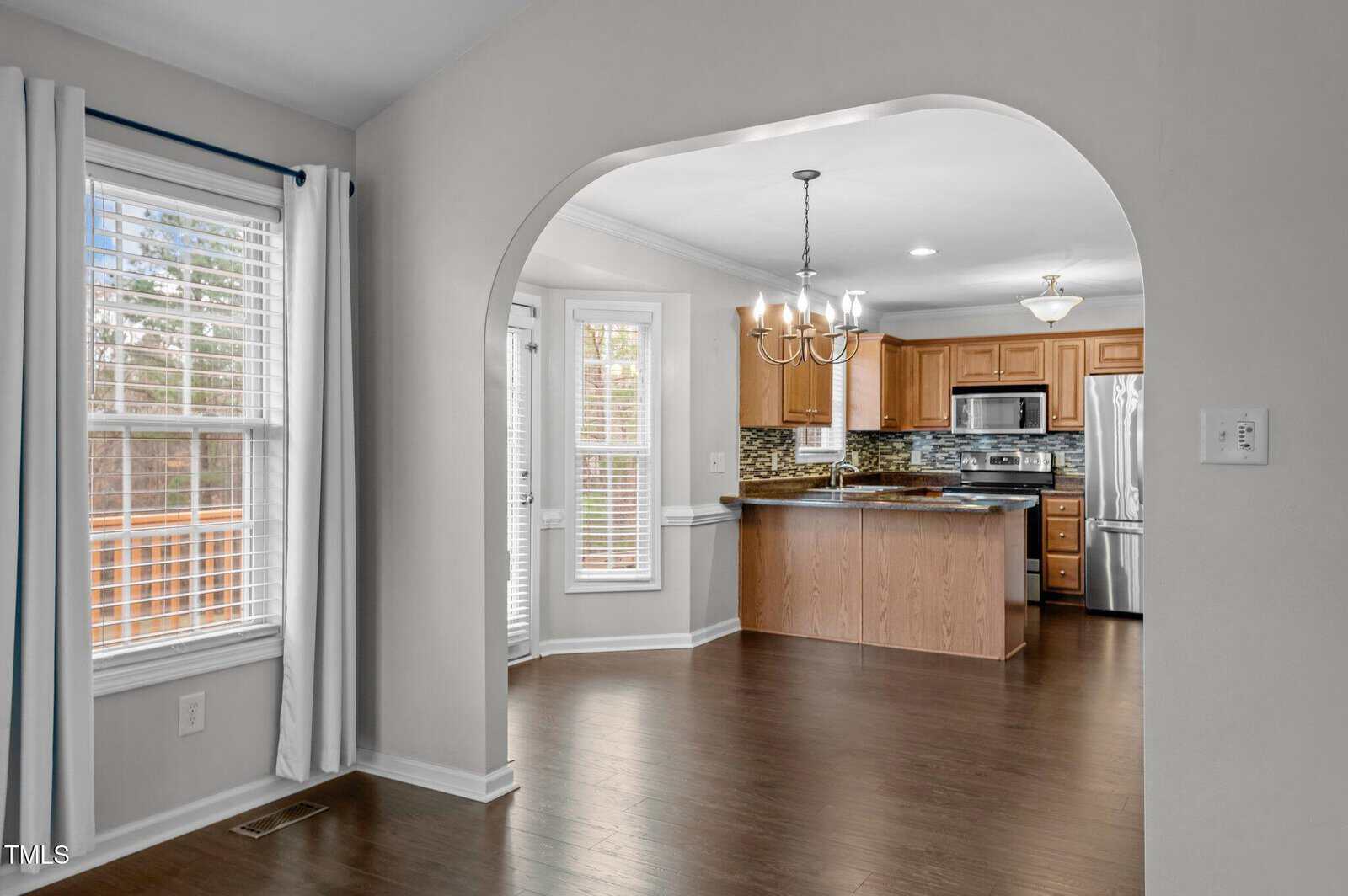 View of a kitchen with wooden cabinets, stainless steel appliances, and an island. Dining area with a chandelier and windows is visible, along with dark hardwood floors and an arched entryway. White walls and curtains are present.