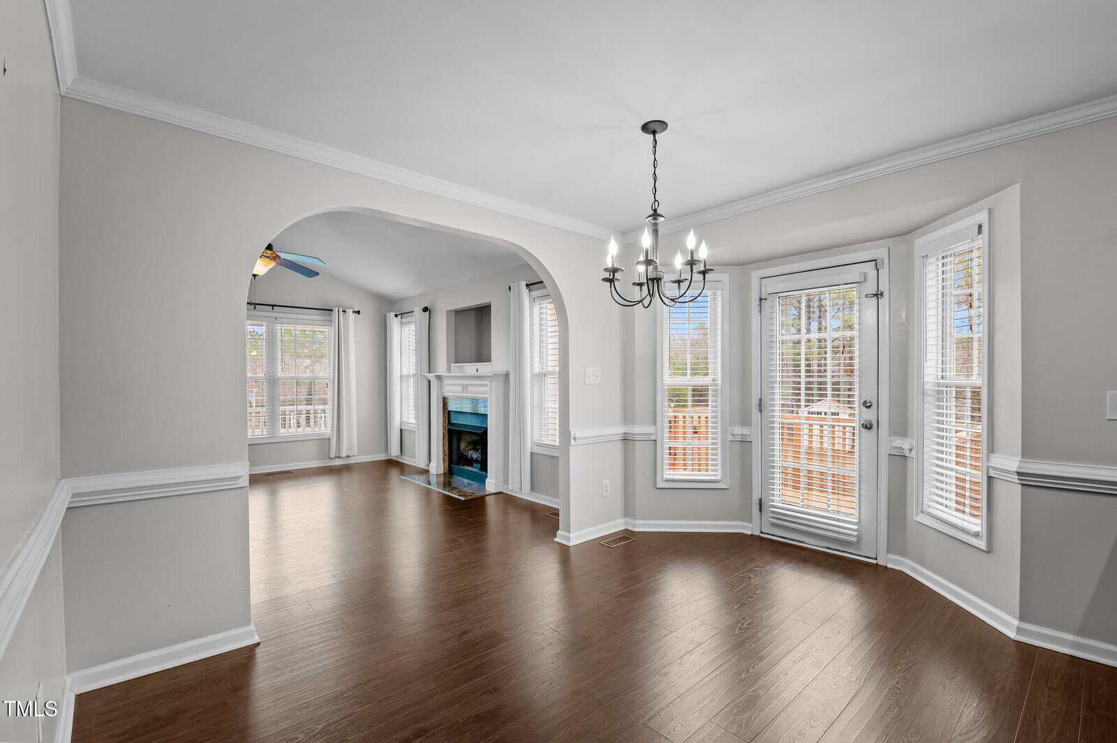 A dining area with wood floors, a chandelier, and French doors next to bay windows. An arched opening leads to a living room with a fireplace and built-in shelves. Walls are light gray, and white trim is visible throughout.