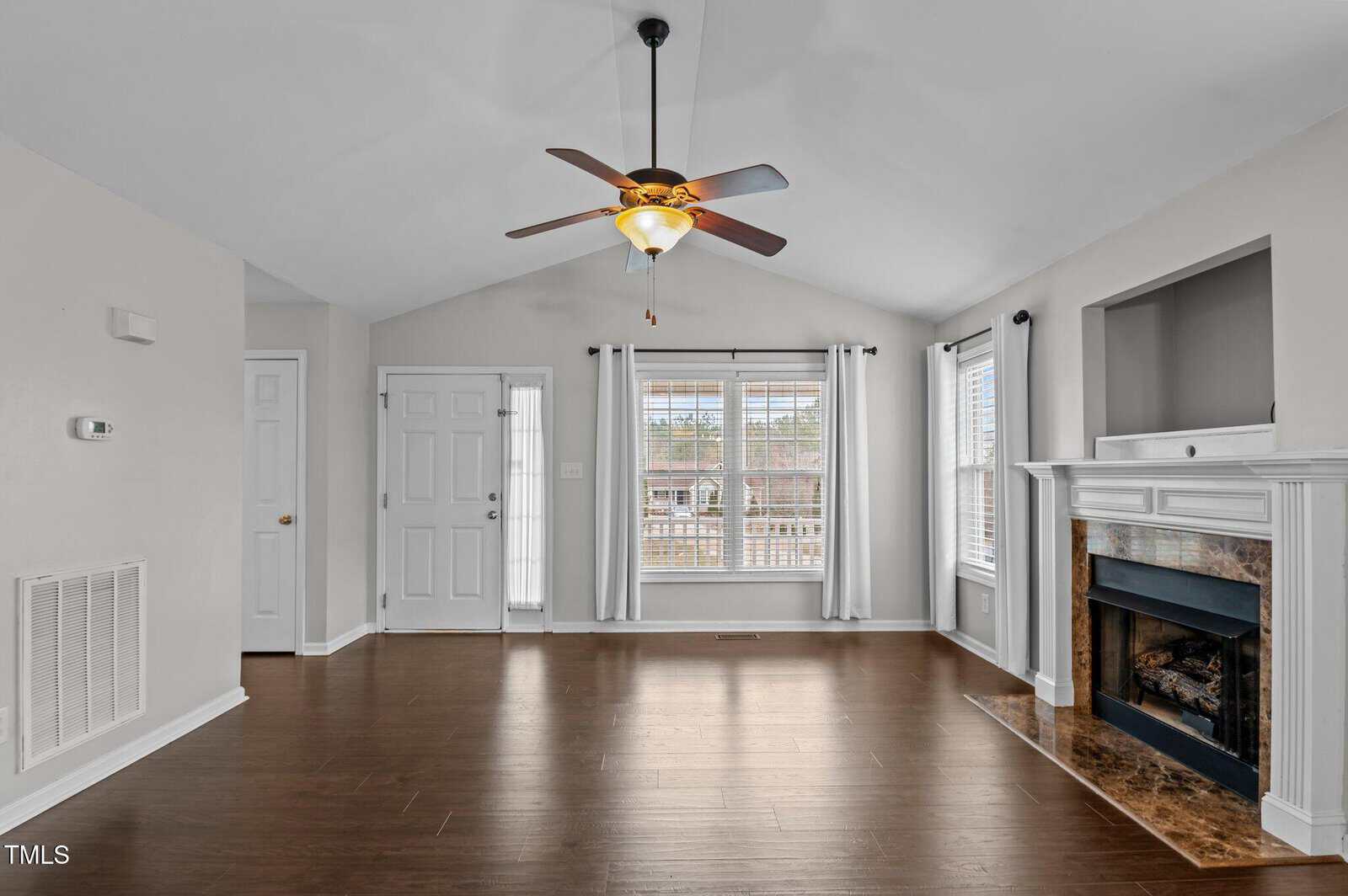 A living room with dark wood floors, a ceiling fan with light, a white front door, large windows with white curtains, and a fireplace with a mantel. The walls are painted light gray.