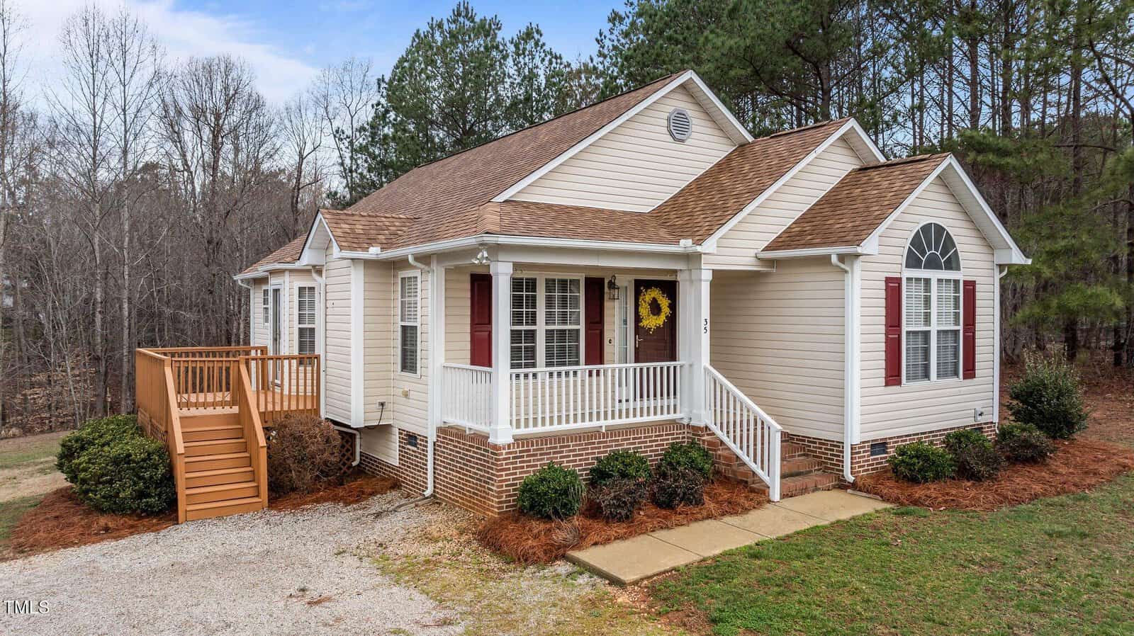 A single-story beige house with a brown roof, white trim, and red shutters. There is a front porch with white railings, a side deck with wooden stairs, and the house is surrounded by trees and pine straw landscaping.