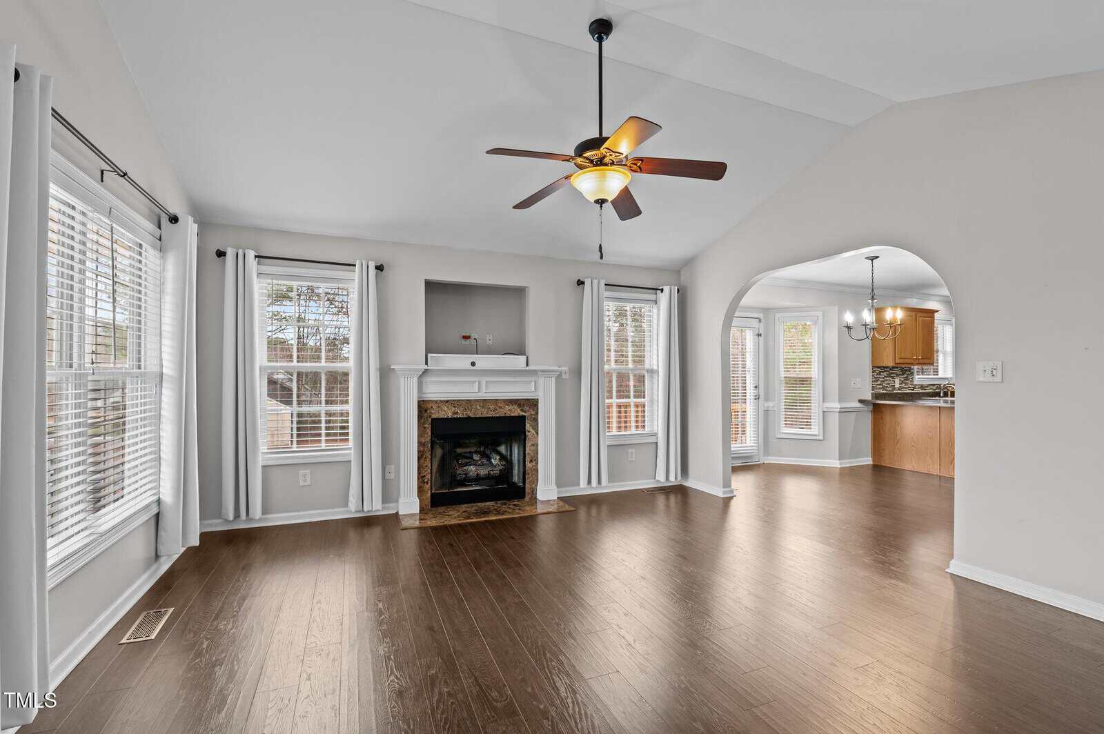 A bright living room with dark hardwood floors, a ceiling fan with lights, a fireplace, and three windows with white curtains, connected to a kitchen through a large arched doorway.