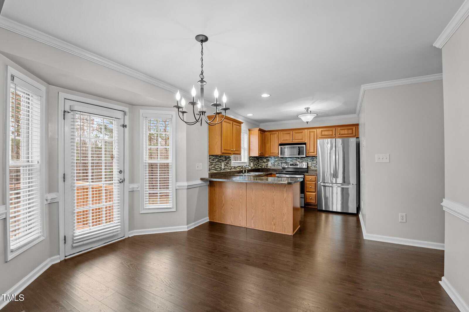 Bright kitchen and dining area with wood floors, stainless steel appliances, wood cabinets, and a tile backsplash. Large windows and glass doors let in natural light. A chandelier hangs over the dining space.