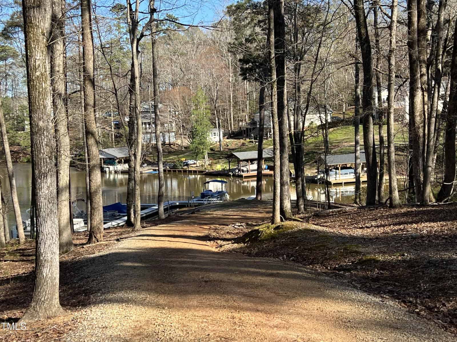 A dirt path leads through trees to a small lake with several docks and boats. Houses are visible on the opposite shore among trees. The scene is sunny with clear skies.