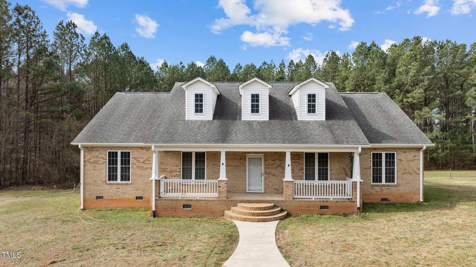 Nestled on 2505 Egypt Mountain Road in Kittrell, a single-story brick house with a gray shingle roof features three dormer windows. The front porch, adorned with white railings and columns, is accessible by a curved concrete pathway. Tall trees frame the home under a partly cloudy sky.