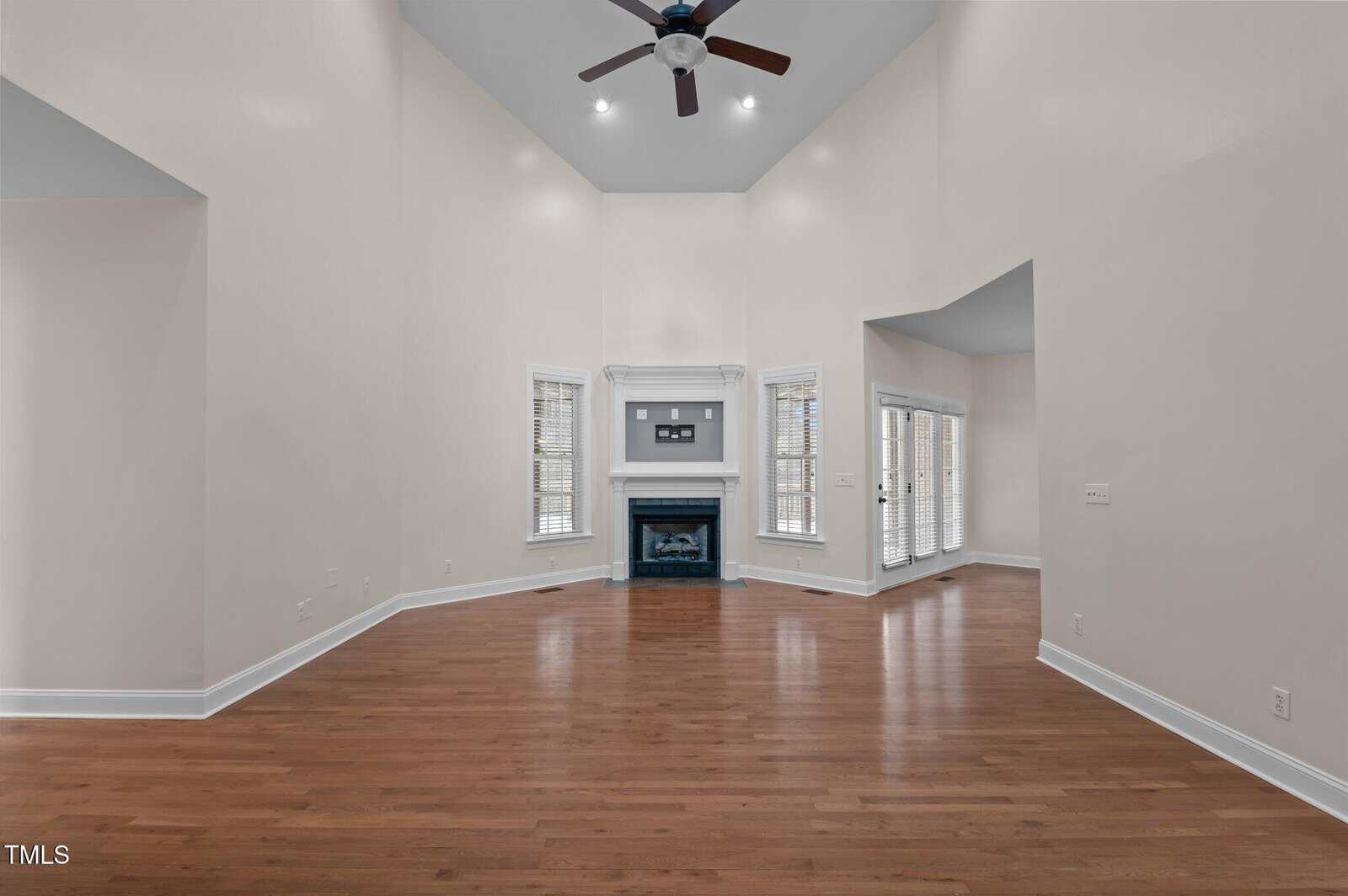 A spacious living room with high vaulted ceilings and a ceiling fan. The room features a fireplace with a mantle, hardwood floors, and two sets of French doors leading to another area. The walls are light-colored.