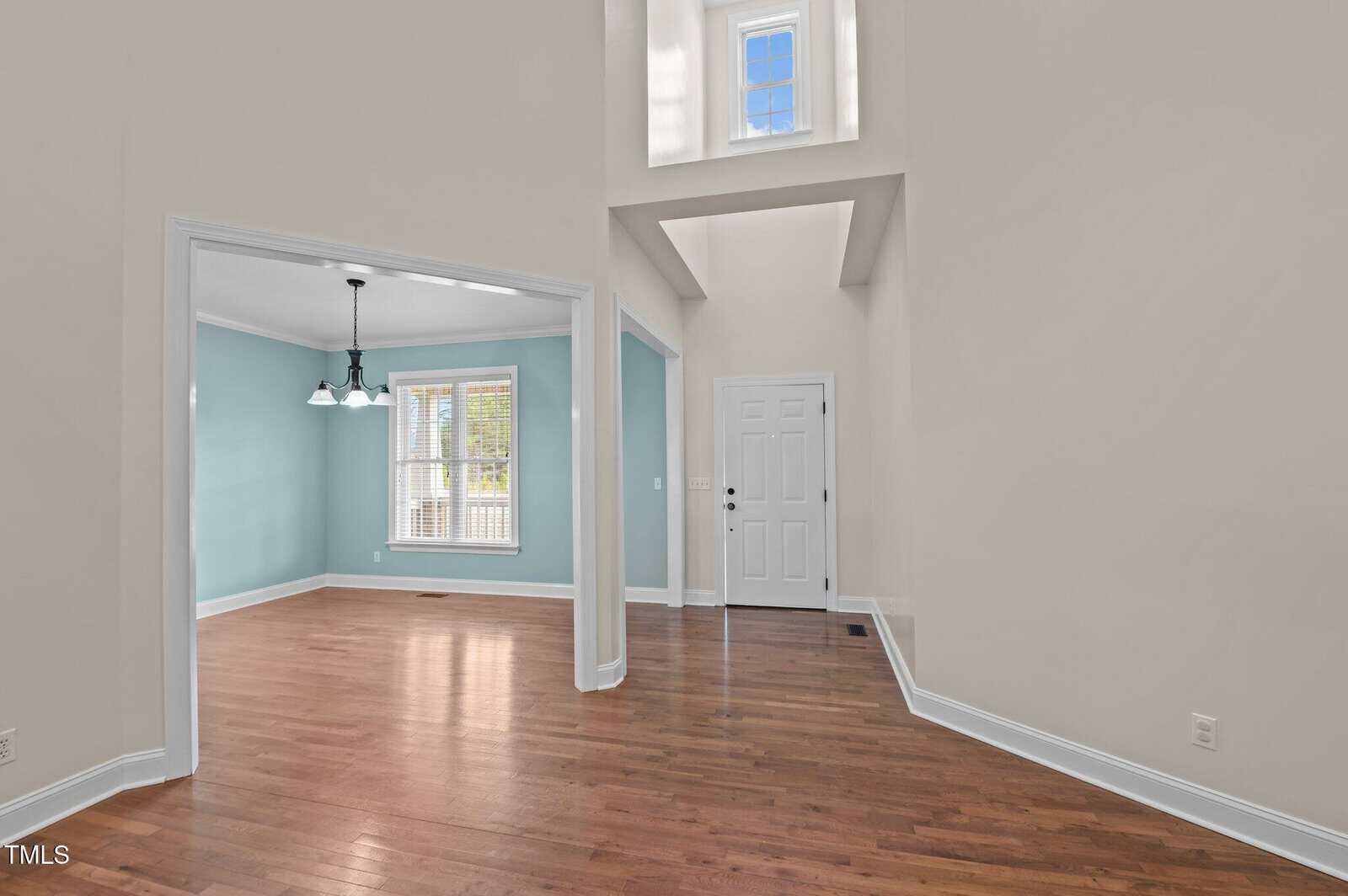 Interior of a house featuring a foyer with a white front door, hardwood floors, and high ceiling. Adjacent room with large window and a white chandelier, painted in a light blue color with white trim. A small window is above the foyer.