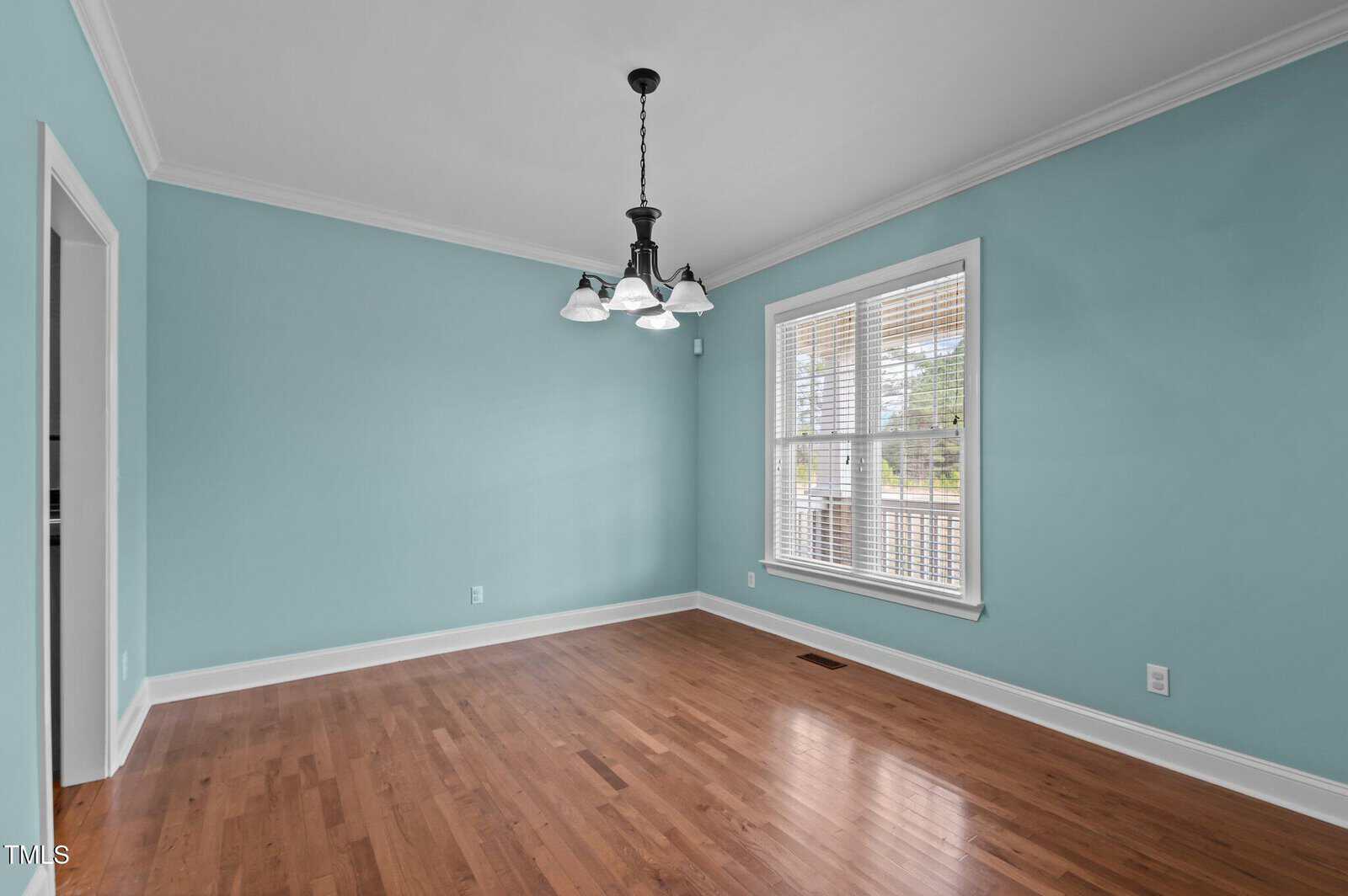 An empty room with light teal walls and a hardwood floor. A window with white blinds is on the right wall, allowing natural light in. A black chandelier with five lights hangs from the white ceiling. A doorway is visible on the left.
