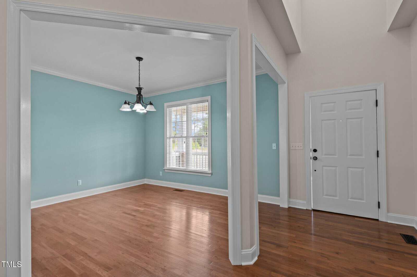 A room with light teal walls and wooden flooring features a window with blinds. There is a three-light chandelier hanging from the ceiling. An open doorway leads to a white door in an adjoining space with similar flooring.