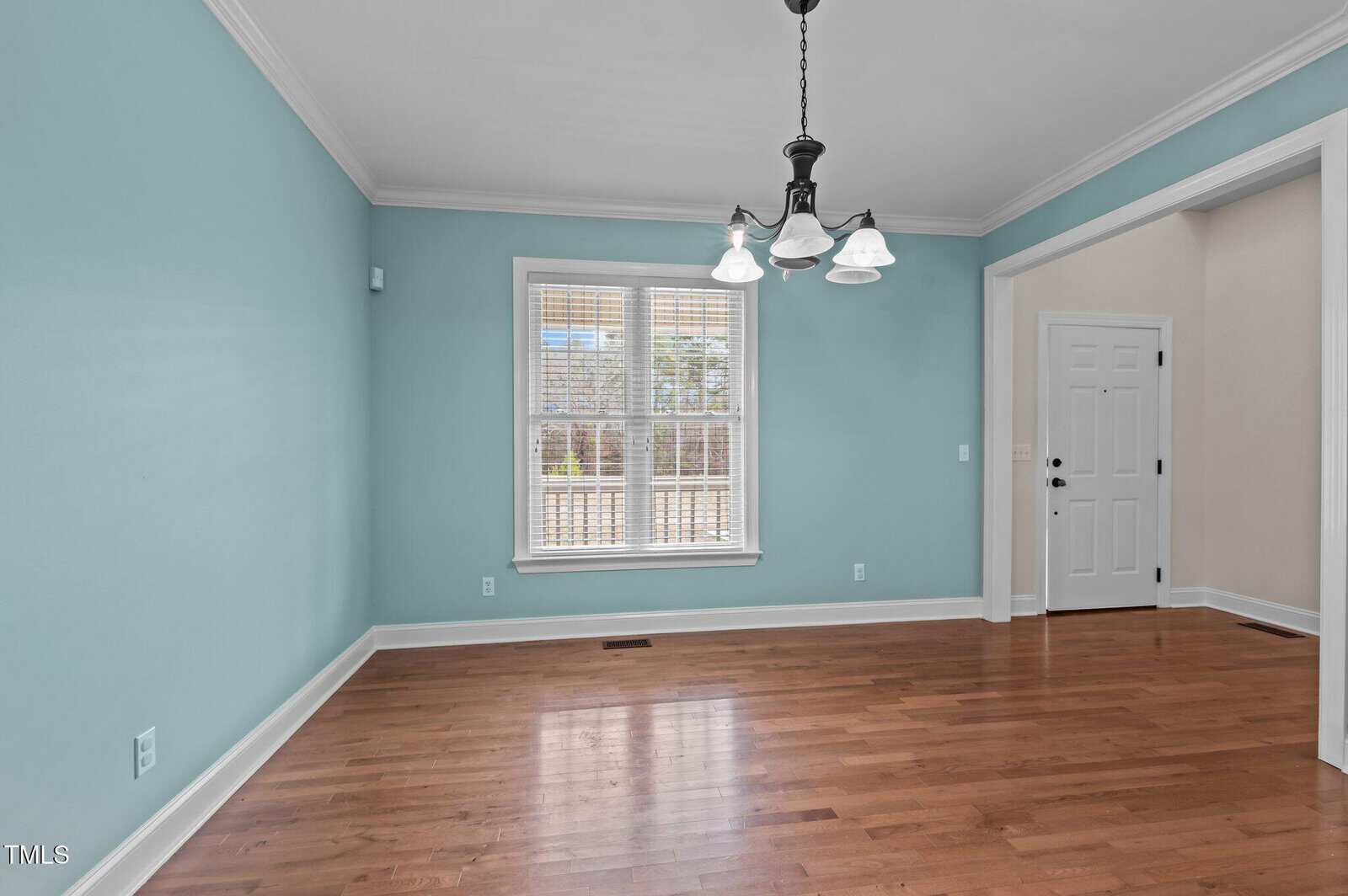 An empty room with light turquoise walls and hardwood floors. A window with blinds allows natural light. A white ceiling features a hanging light fixture. There's a white door in the adjacent hallway.