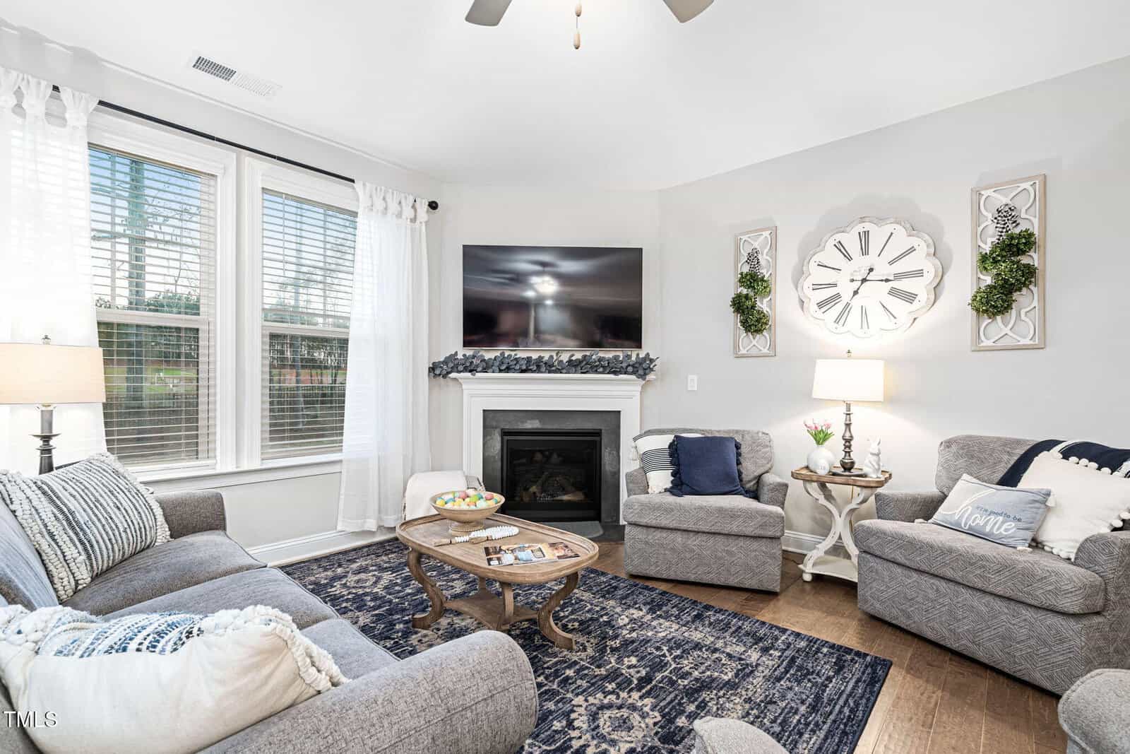 A bright living room with gray sofas, a wooden coffee table, a dark blue patterned rug, a fireplace with a TV above it, and large windows with white curtains letting in natural light.