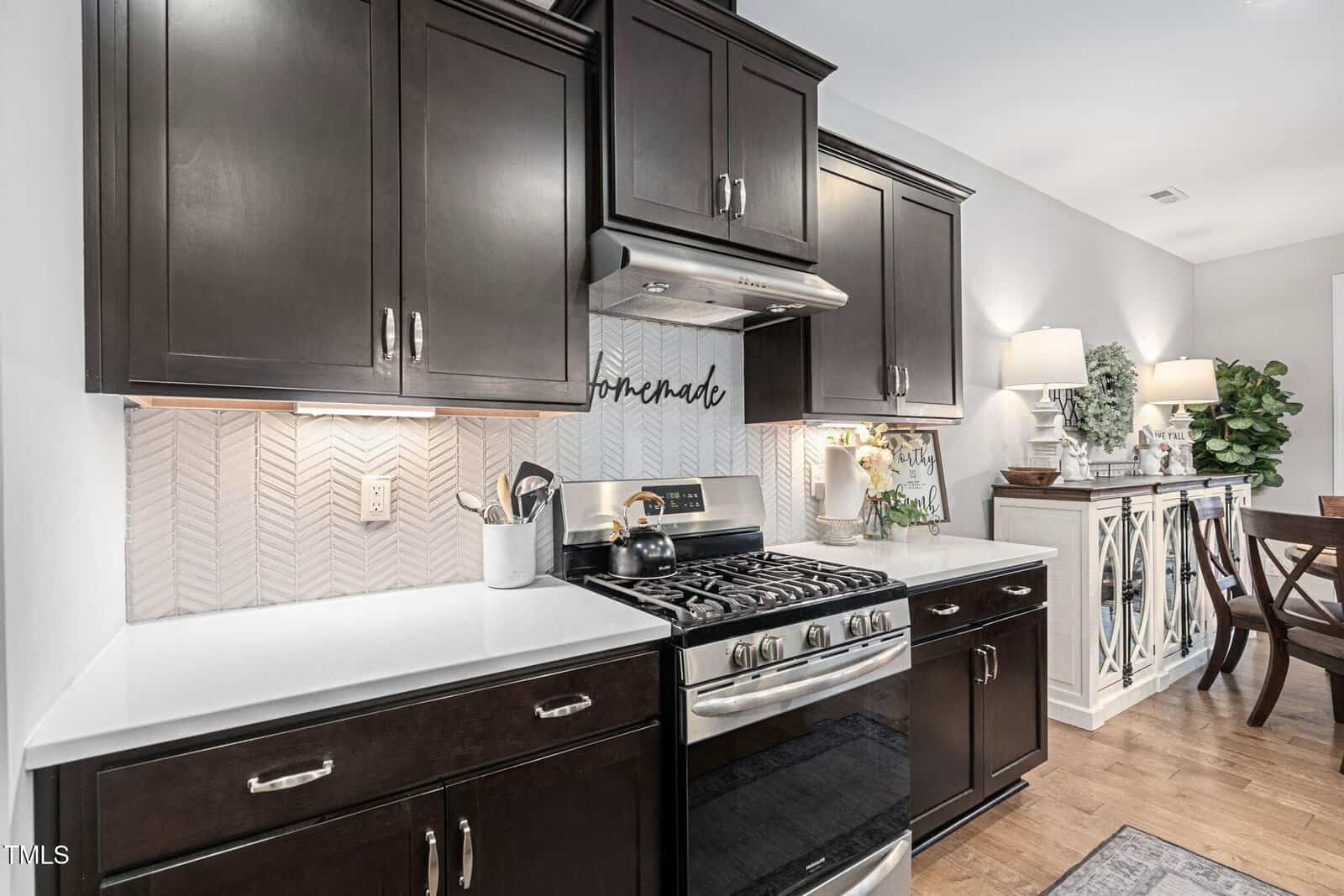 Kitchen with dark cabinets, white countertops, stainless steel gas stove, herringbone backsplash, and a “homemade” sign above the range. Dining area with plants and a white sideboard in the background.