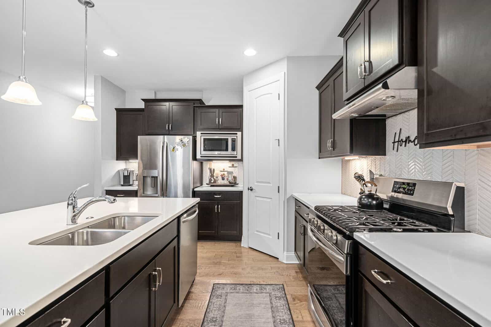 Modern kitchen with dark cabinets, stainless steel appliances, white countertops, a central island with a sink, pendant lights, and light wood flooring. The oven and refrigerator are built-in, and a small rug is on the floor.