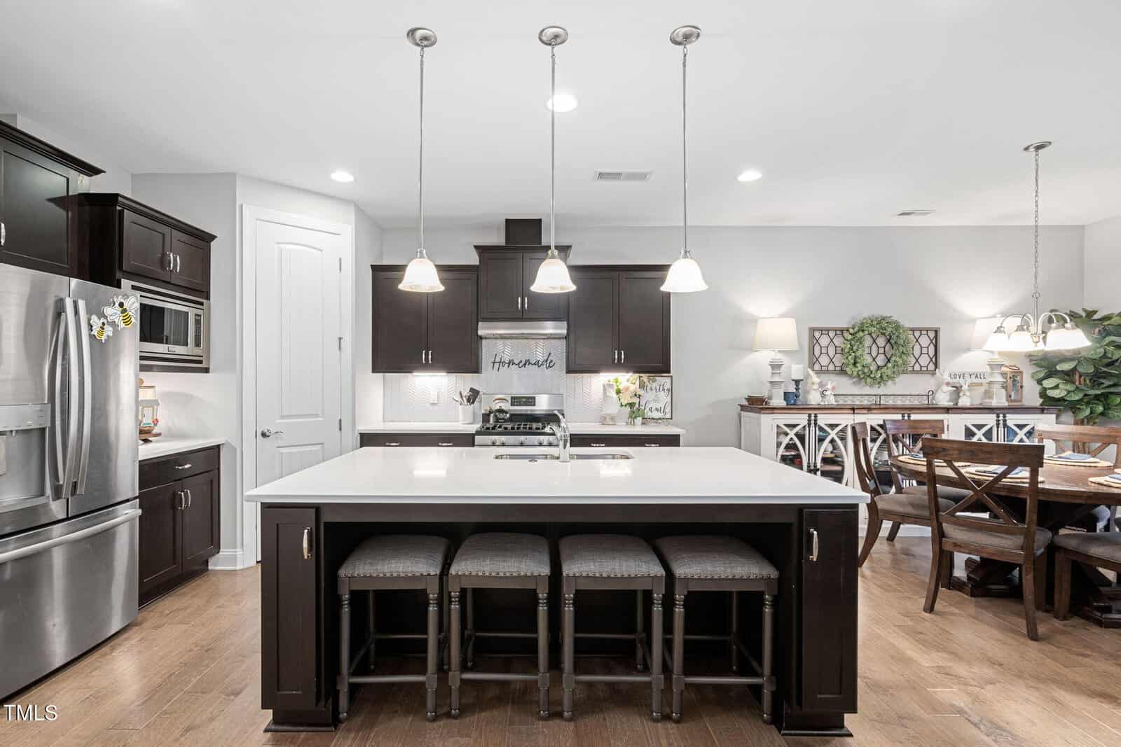 Modern kitchen with a large island, three upholstered bar stools, dark cabinets, stainless steel appliances, pendant lights, and a dining area with a wooden table and chairs in the background.