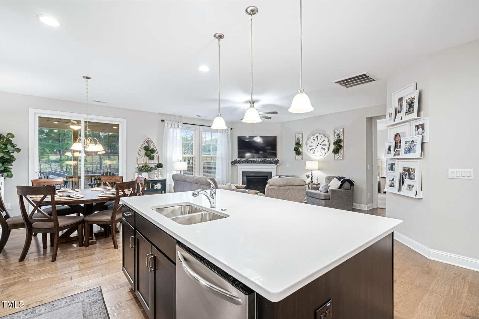Open-concept kitchen and living area with a white island, stainless steel sink, and dishwasher. Dining table with chairs to the left and a living room with sofas, TV, and wall decor in the background.