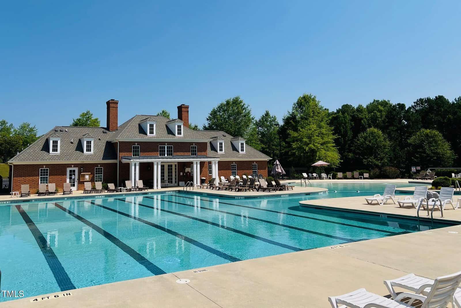 A large outdoor swimming pool with lap lanes, surrounded by lounge chairs and umbrellas, is in front of a two-story brick clubhouse with tall windows and chimneys. Trees and clear blue sky are in the background.