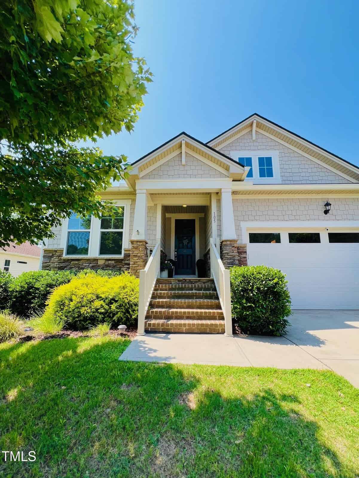A two-story suburban house with beige siding, stone accents, and a white garage. Brick steps lead to a covered front porch with white railings. A green lawn and tree are visible in the foreground.
