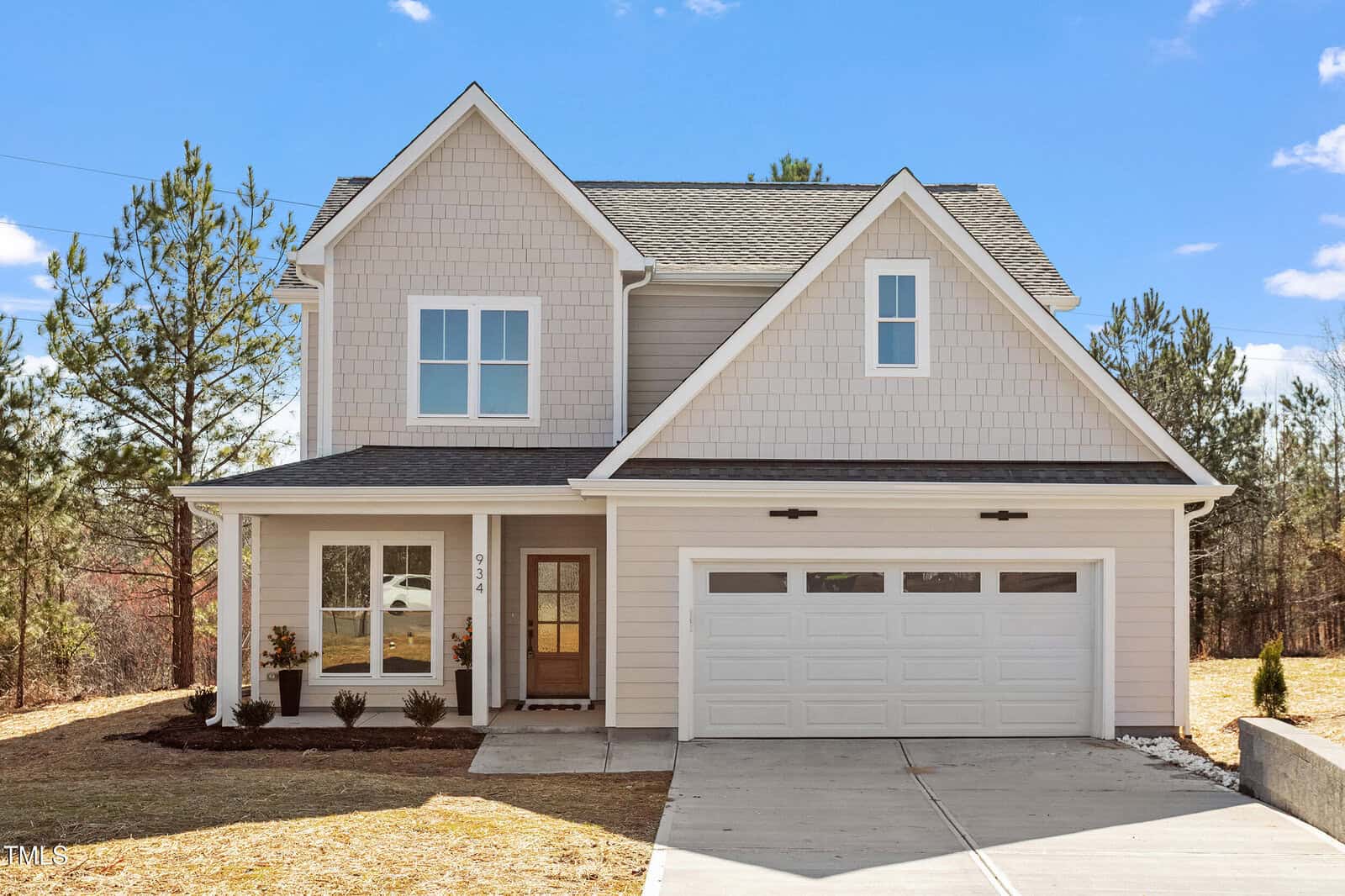 Nestled on 934 Woodland Road in Creedmoor, this charming two-story house boasts light gray siding and a dark gray roof. It features a welcoming front porch with white columns, a double garage door, and three front-facing windows. A driveway and small lawn complement the serene backdrop of trees.