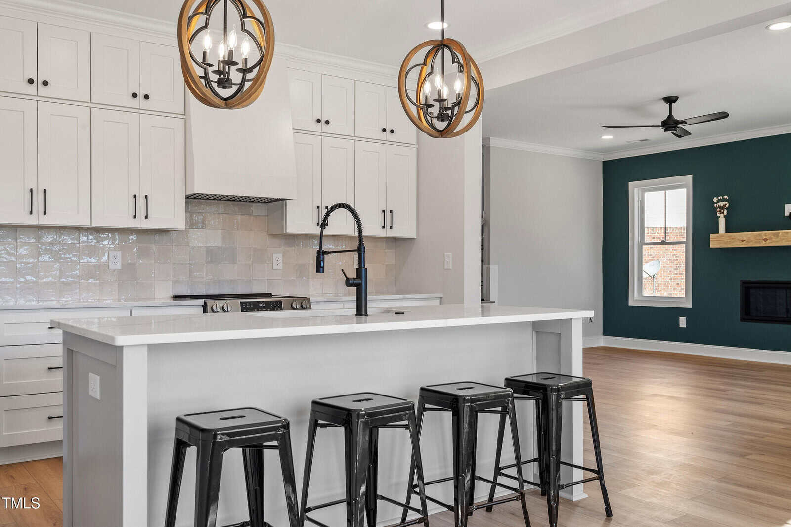 Modern kitchen interior featuring a white island with four black stools. There are white cabinets, a tiled backsplash, and hanging pendant lights with circular wood accents. An adjacent living area with a dark green accent wall and ceiling fan is visible.