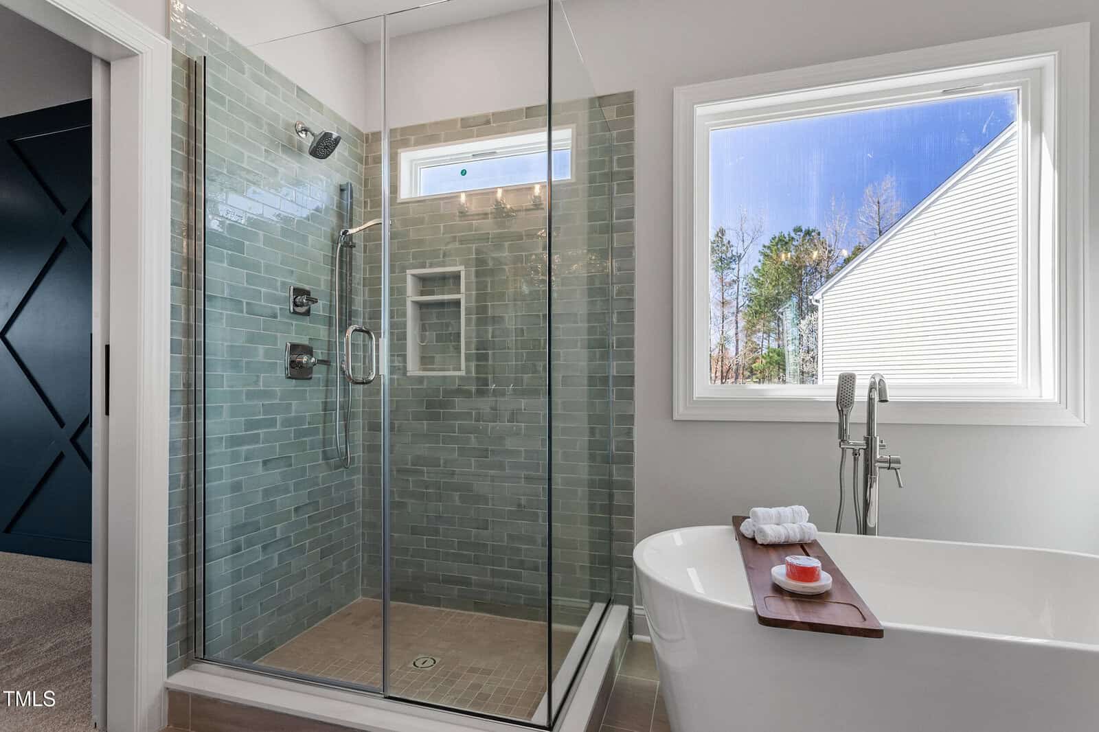 A modern bathroom features a glass-enclosed shower with green subway tiles, a small window, and chrome fixtures. Nearby is a white freestanding bathtub with a towel and bottle on the edge, positioned next to a large window showing an outdoor view.