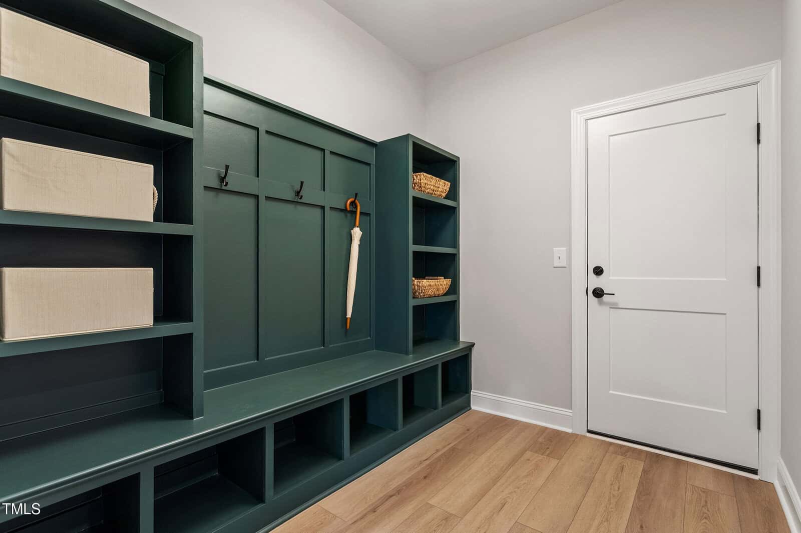 A mudroom with dark green built-in shelves and a bench. The shelves contain beige and wicker storage boxes. A white umbrella hangs on a black hook. The walls are light gray, and the flooring is light wood. A white door with a black handle is on the right.