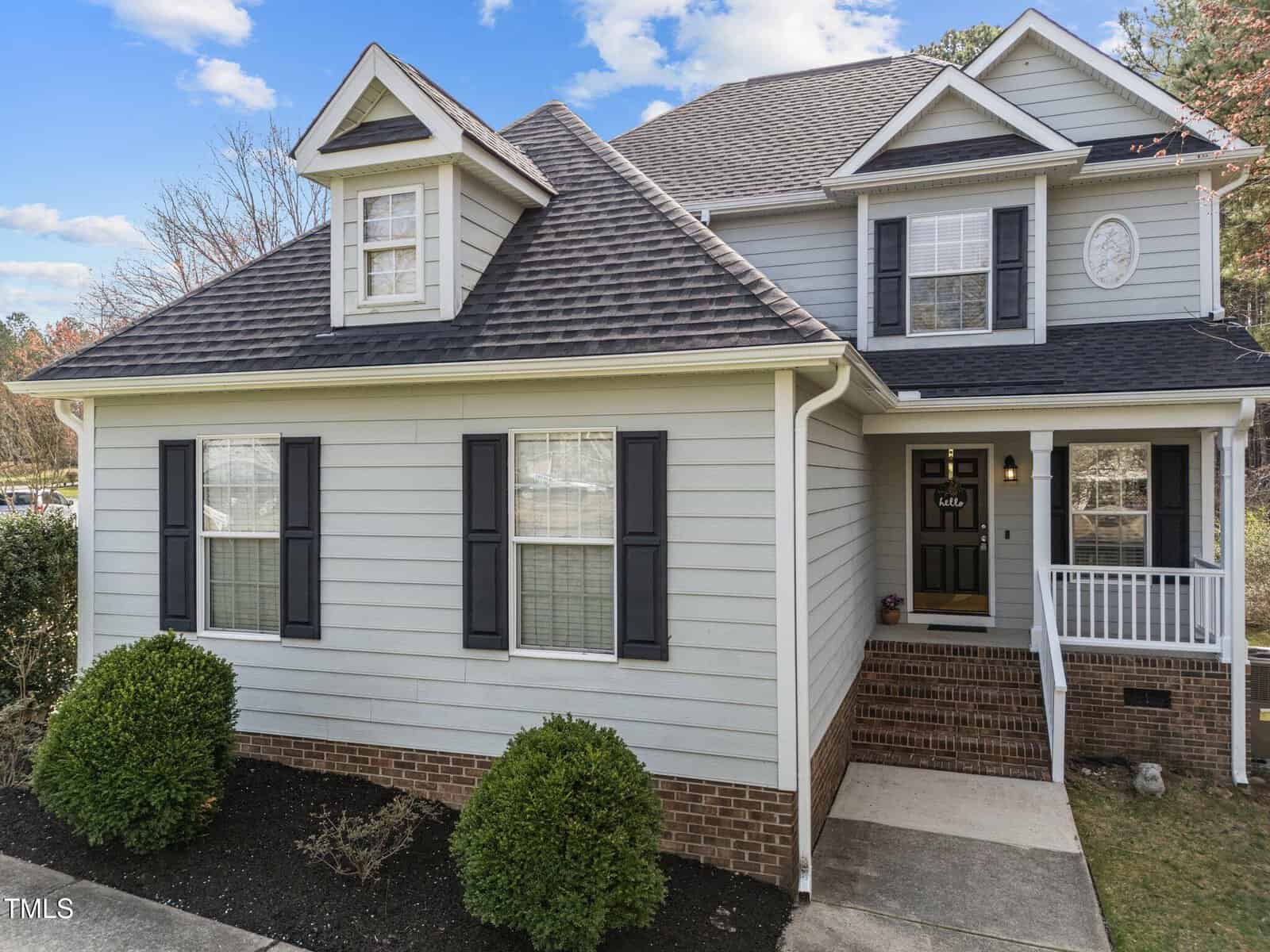 A two-story house with light gray siding, dark shutters, and a brick foundation. There are trimmed bushes in front, a small covered porch with white railings, and a dormer window under a blue sky.