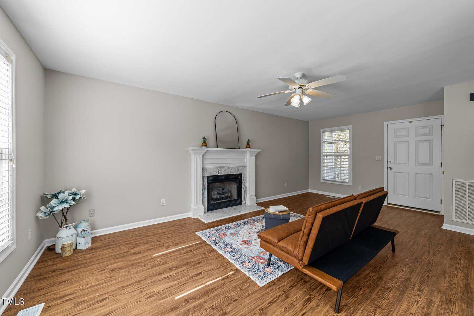 A living room with wood flooring, a white fireplace, a ceiling fan with lights, a black and wood futon, a patterned rug, and large windows with blinds. The walls are light gray and the room is minimally decorated.