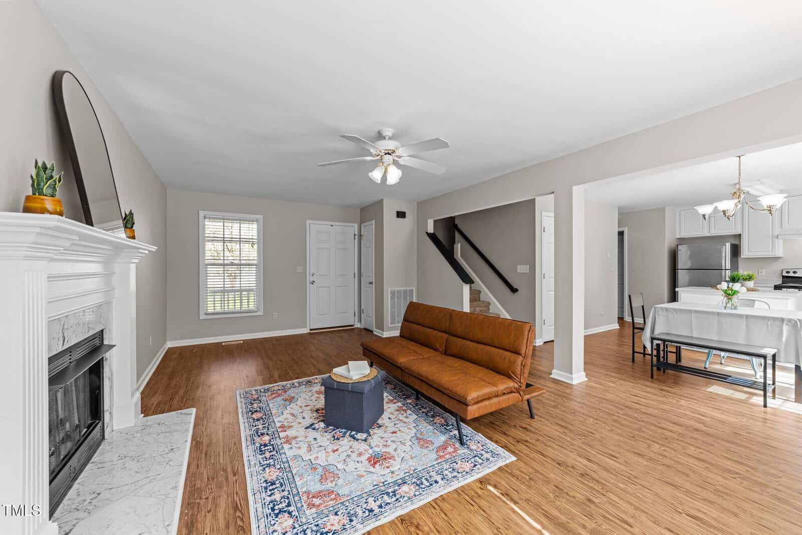 A modern living room with wood flooring, a brown leather sofa, a small ottoman on a patterned rug, a white fireplace, and an open view of the dining area and kitchen with stainless steel appliances.