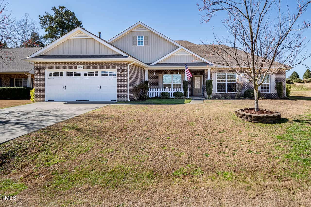 Located at 1176 Andrews Court in Creedmoor, this single-story brick house features a two-car garage and a lush front yard with a small tree on the right. An American flag waves proudly near the entrance, all beneath a clear blue sky.