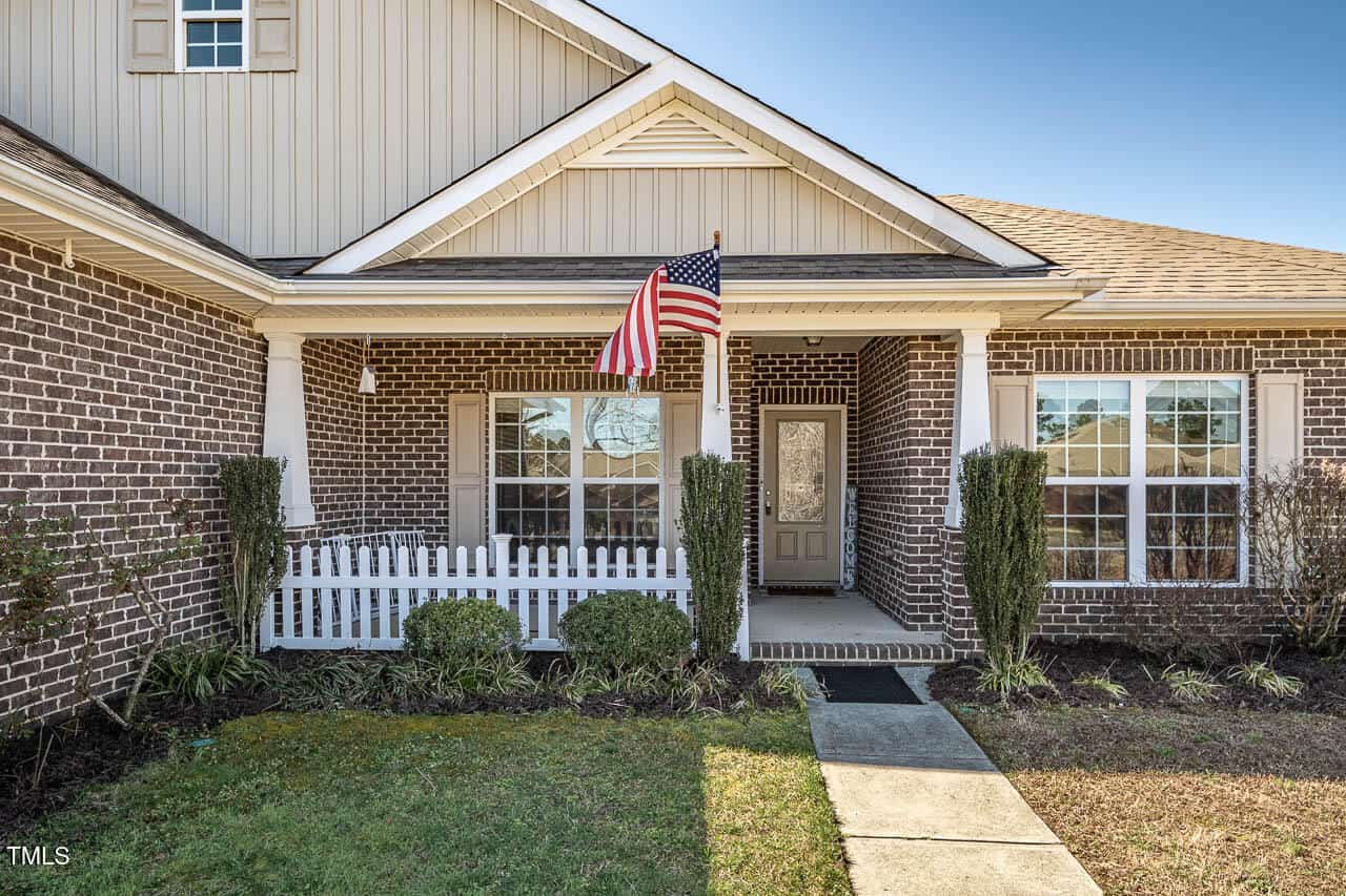 Single-story brick house with a front porch featuring a white fence and an American flag. There are two large windows on either side of the entrance, with trimmed shrubbery and a pathway leading to the front door.