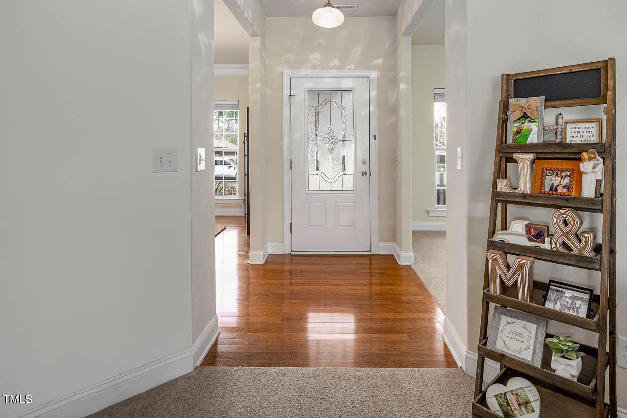A hallway with wooden flooring and light-colored walls leads to a white front door with decorative glass. A wooden ladder shelf on the right holds framed photos and decorative items. Natural light filters through windows on either side.