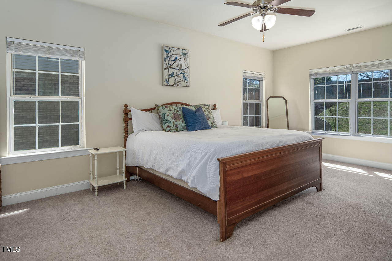 A bedroom with a wooden bed frame and white bedding. There are several decorative pillows on the bed. The room has light-colored walls, a ceiling fan with lights, three windows with blinds, and a small side table next to the bed.