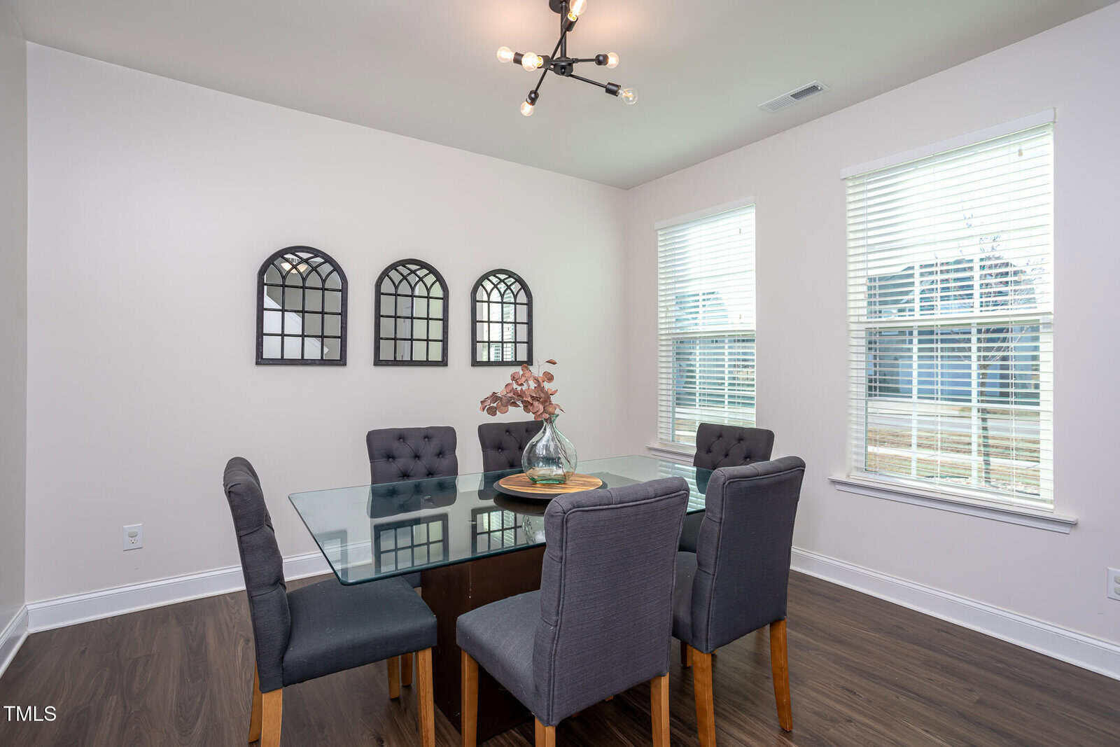 A dining room with a glass table, six gray upholstered chairs, a modern black light fixture, and three arched wall mirrors. Two large windows with blinds let in natural light. A vase with dried flowers is on the table.