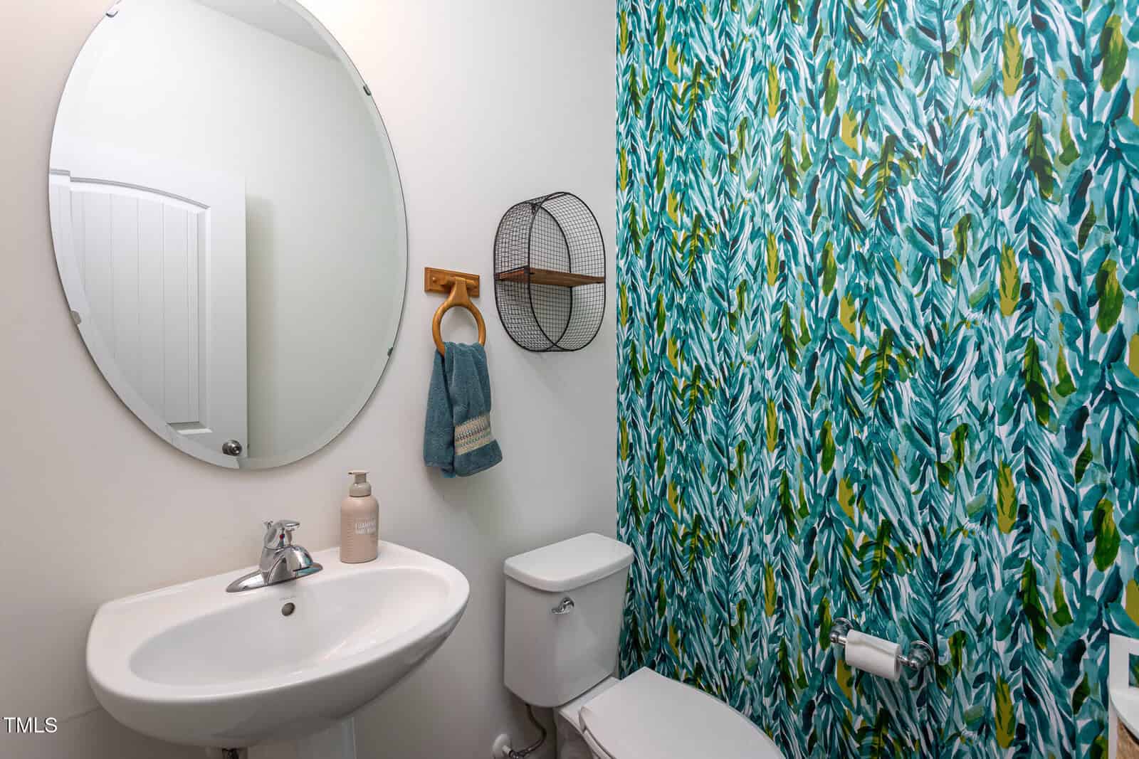 A small bathroom with a white sink, soap dispenser, round mirror, white toilet, and a blue-green leafy patterned accent wall. A towel hangs on a wooden holder next to a metal shelf above the toilet.